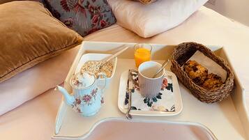 A breakfast tray on a bed with a teapot, mug, cereal, orange juice, and a basket of cookies.
