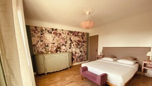 Ein Schlafzimmer mit Holzboden, einem Bett mit weißer Bettwäsche, einer pinkfarbenen Bank, floralem Wandpapier, einem grünen Schrank und einer Deckenleuchte.