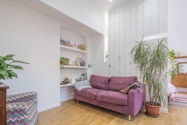 A living room with a purple sofa, wooden flooring, a large potted plant, and a white wall with shelves containing books and decorative items. - Central Cozy Nest With Parking Bogliasco (Bogliasco)