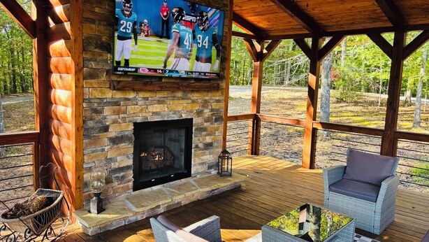 Un espacio al aire libre techado con chimenea de piedra, muebles de mimbre y un televisor en la pared que muestra un partido de fútbol.