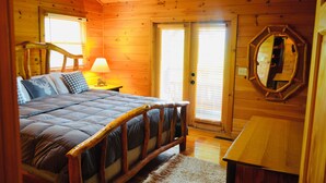 Un dormitorio de cabaña de madera con una cama grande, una mesita de noche con lámpara, un espejo y una mesa de madera.