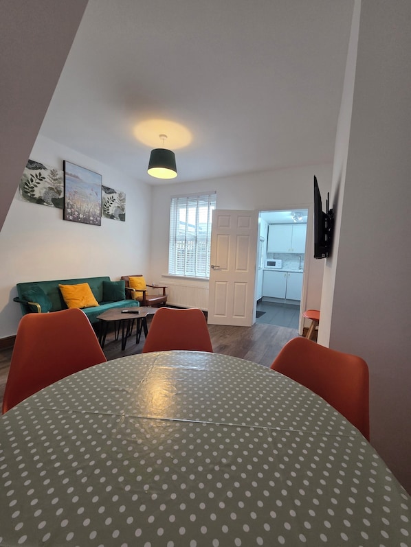 Un salon avec un canapé vert, des fauteuils orange, une table à manger recouverte d’une nappe à pois, un tableau accroché au mur et un téléviseur fixé au mur.