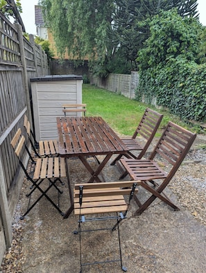 Un patio con mesa y sillas de madera, un cobertizo pequeño y un jardín con cerda y árboles.