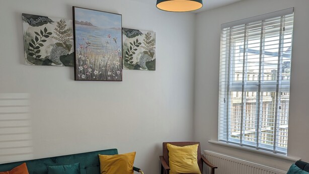 A living room with a green sofa, yellow cushions, a wooden coffee table, and a brown armchair. There are two framed botanical prints on the wall and a window with blinds.