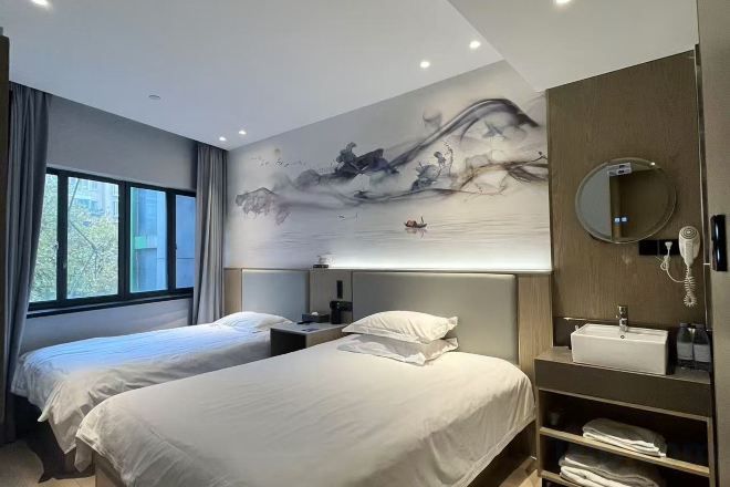 A modern hotel room with two beds, a large artwork on the wall, a bedside table with a lamp, and a built-in shelf with towels.
