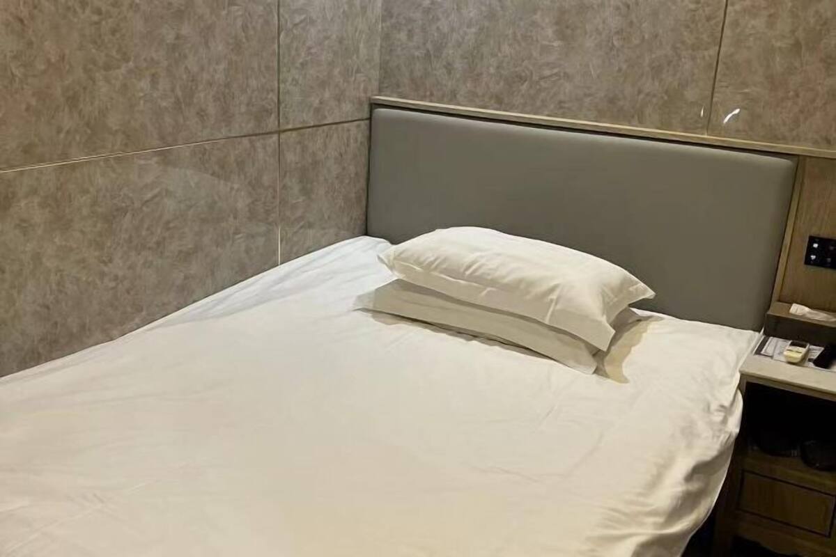 A neatly made bed with a grey headboard, a bedside table with a lamp, and a wall-mounted control panel.