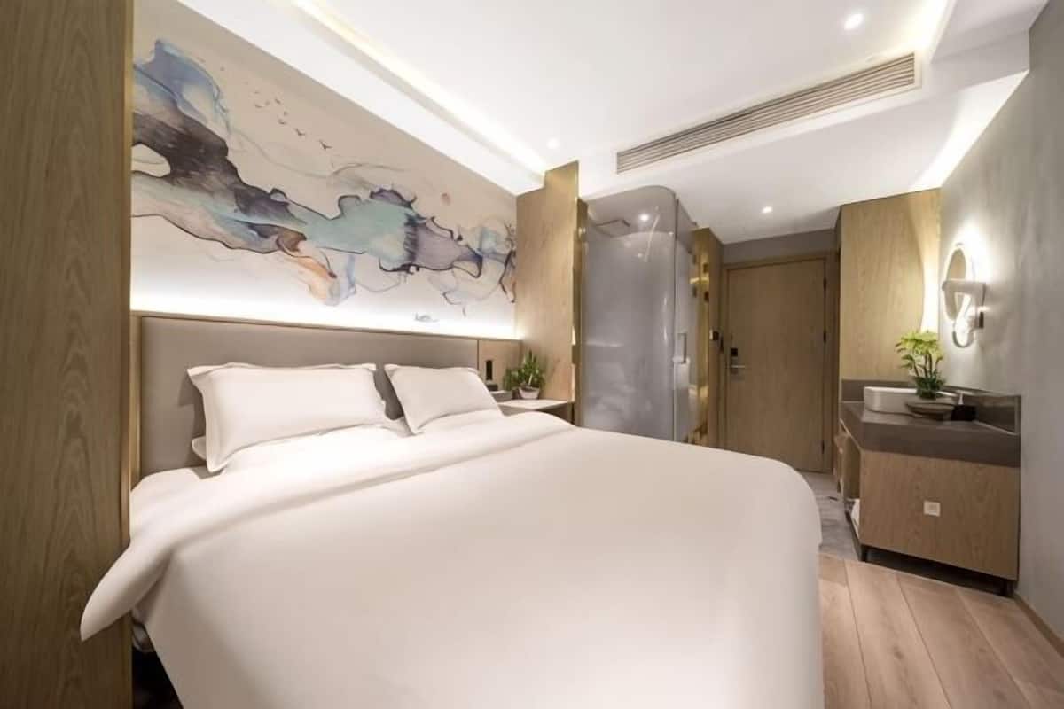 A modern hotel room with a large bed, a wall-mounted artwork, a built-in wardrobe, and a bathroom with a mirror and sink.