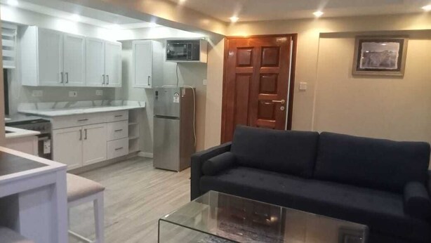 A compact living space with a kitchenette, a glass coffee table, a dark sofa, and a wooden door. - Kenzi Suites (Kisii)