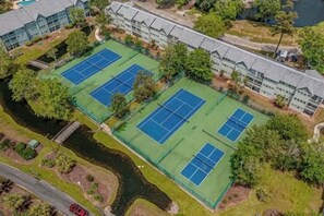 An aerial view of a hotel with multiple tennis courts, a parking area, and a body of water nearby.
