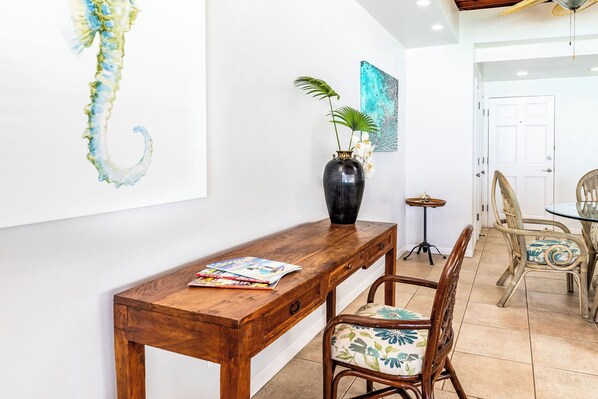 Un escritorio de madera con un florero y una revista, una silla con un cojín floral, y una pared con un cuadro de caballito de mar y un cuadro abstracto azul.