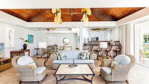 A spacious living area with wicker furniture, a wooden ceiling, and a view to the kitchen.