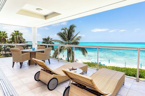 A balcony with wicker furniture, a table, and a view of the ocean and palm trees.