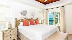 A bedroom with a large bed, a nightstand, a bench, and a view of the ocean.