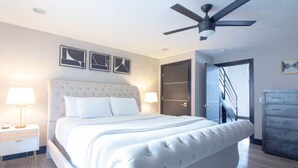 Une chambre à coucher avec un grand lit, deux tables de chevet avec des lampes, un ventilateur de plafond et une porte donnant sur une autre pièce.
