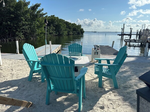 A beachside seating area with turquoise Adirondack chairs and a table, overlooking calm waters and a dock with boats.