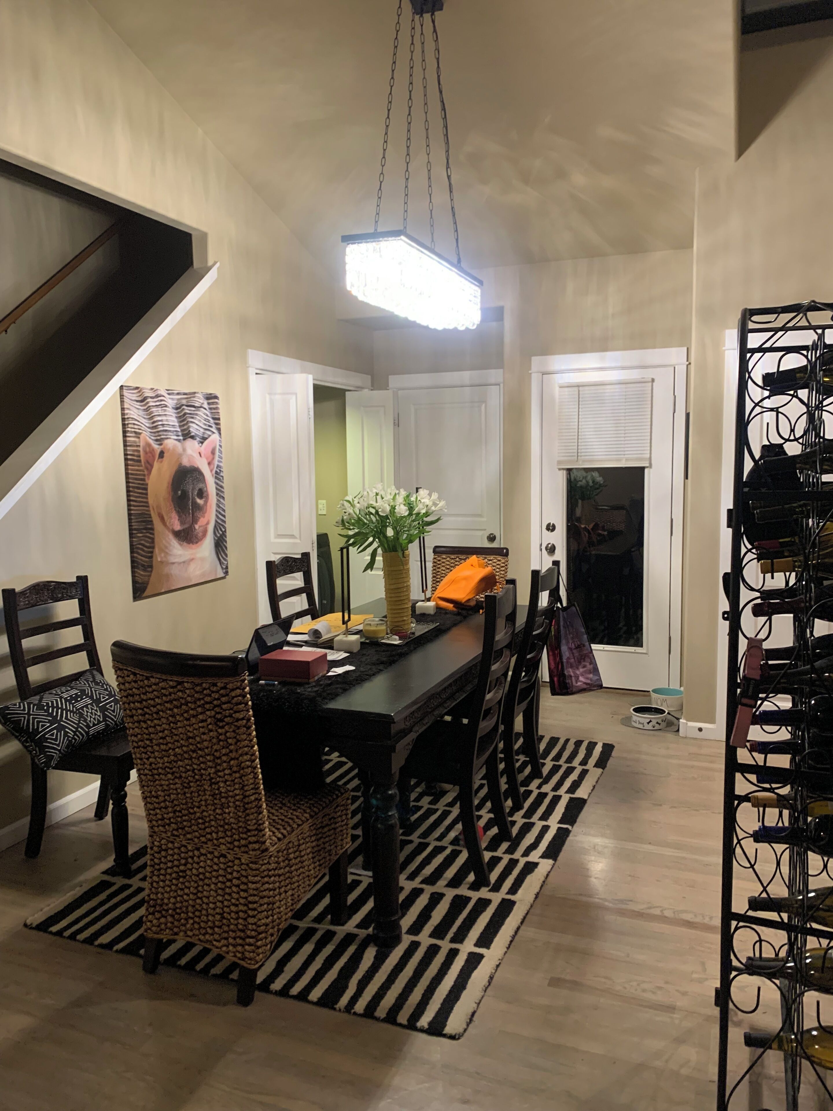 A dining area with a black table, chairs, and a hanging light fixture. There is a painting on the wall and a wine rack to the side.