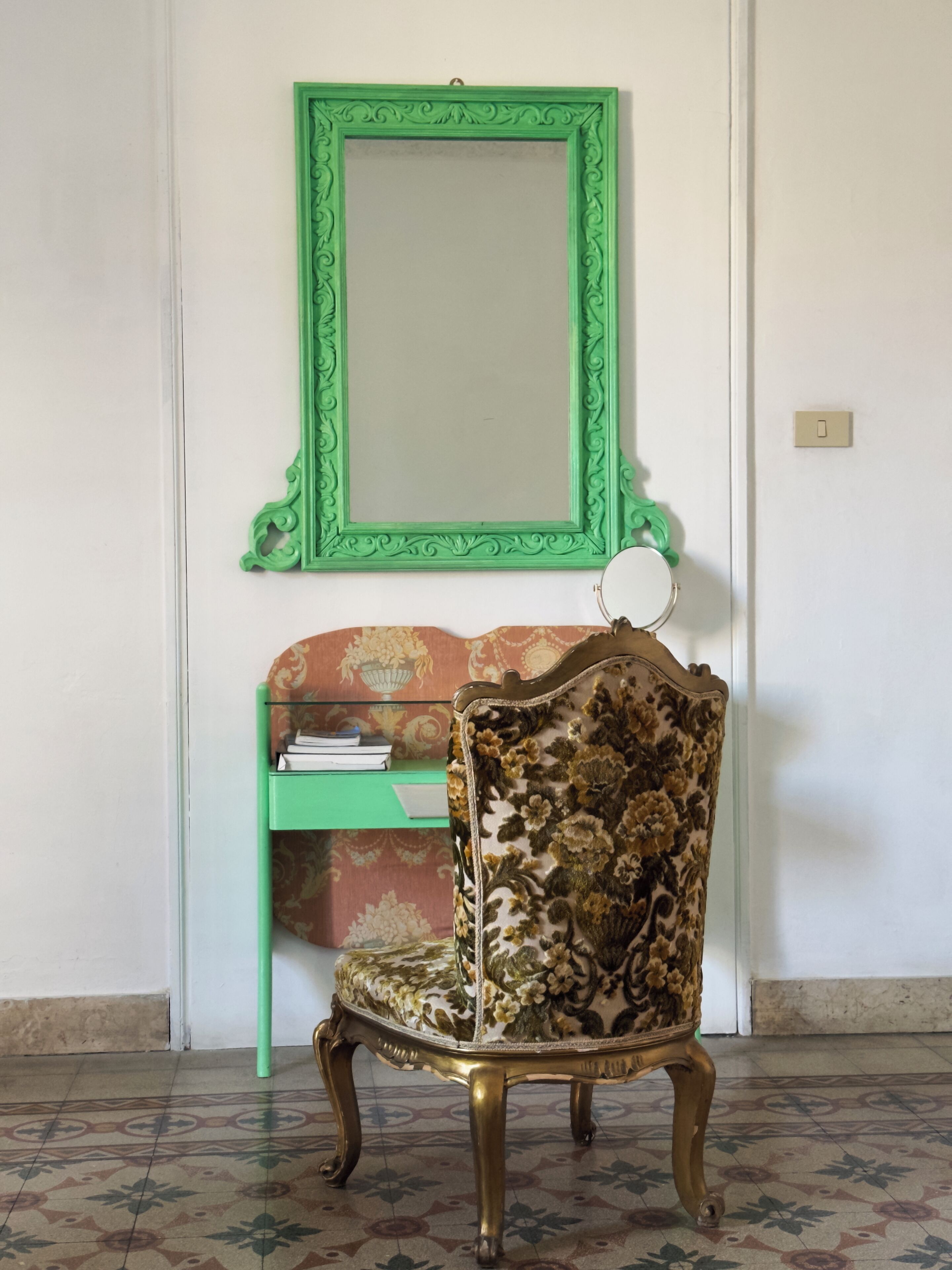 Una sedia con motivo floreale, uno specchio con cornice verde e una mensola verde con libri.