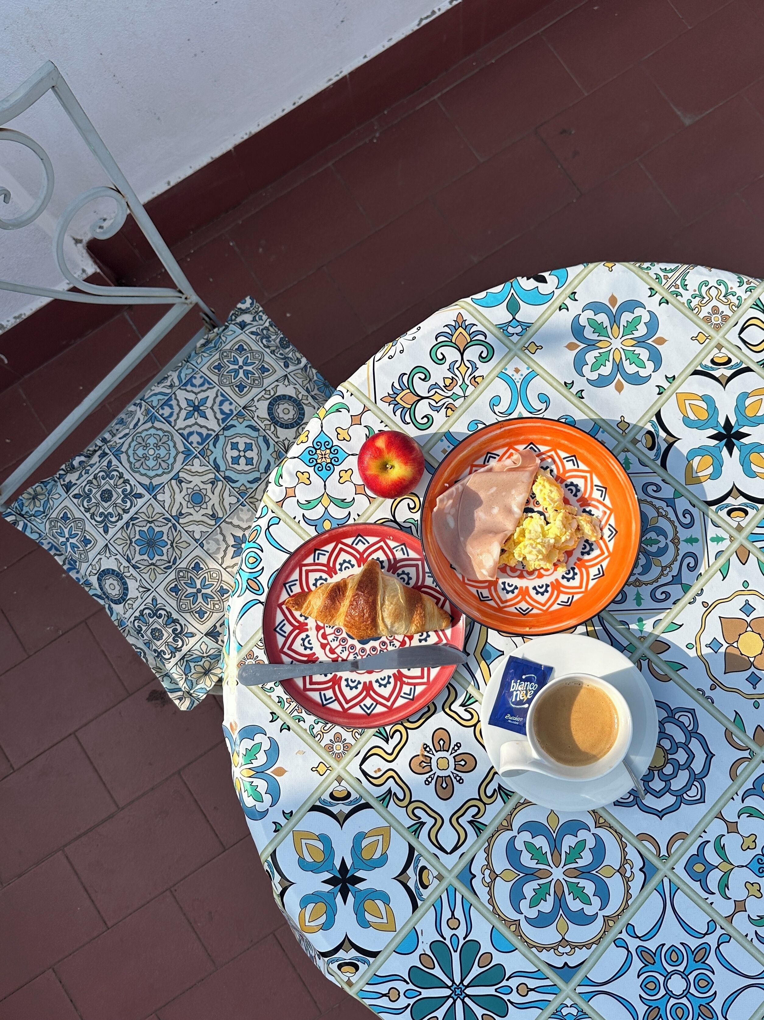Un tavolo rotondo con una tovaglia fantasia, un piatto con un croissant, una ciotola di uova strapazzate, una mela e una tazza di caffè.