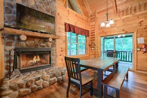 A cozy cabin interior with a stone fireplace, wooden walls, a dining area, and a large window offering a view of the outdoors.