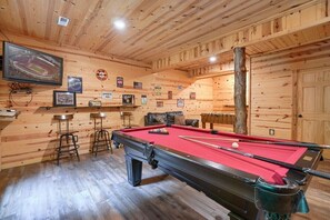 A pool table with a red felt surface, surrounded by wooden walls with various signs and a mounted television. - Twin Creeks | Hocking Hills (Logan)
