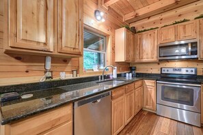 A wooden cabin kitchen with a granite countertop, stainless steel appliances, and a window with a view of greenery.