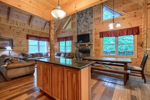 A cozy cabin interior with a wooden interior, a stone fireplace, a dining area, and a large window overlooking greenery.