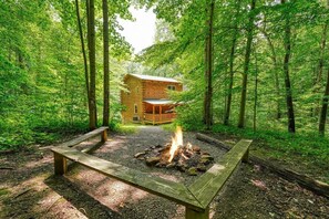 A cabin surrounded by a forest with a fire pit and benches.