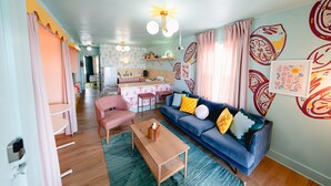 A living room with a blue sofa, pink chairs, a wooden coffee table, and a kitchen in the background.