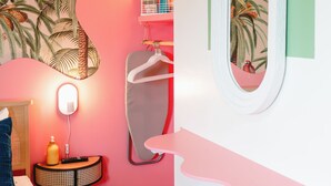 A room with a vibrant pink and white color scheme, a large tropical-themed wall mural, a round mirror with a green frame, and a bed with striped bedding.