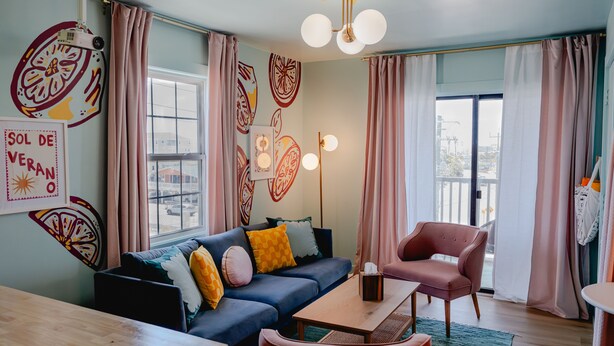A modern living room with a blue sofa, a wooden coffee table, and a pink armchair. There are large windows with curtains, a ceiling light fixture, and wall art.
