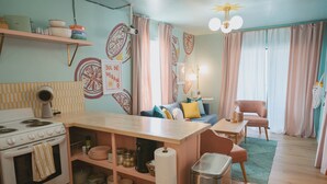 A cozy kitchen with a stove, shelves with dishes, a dining area with a table and chairs, and a living room with a sofa and curtains.