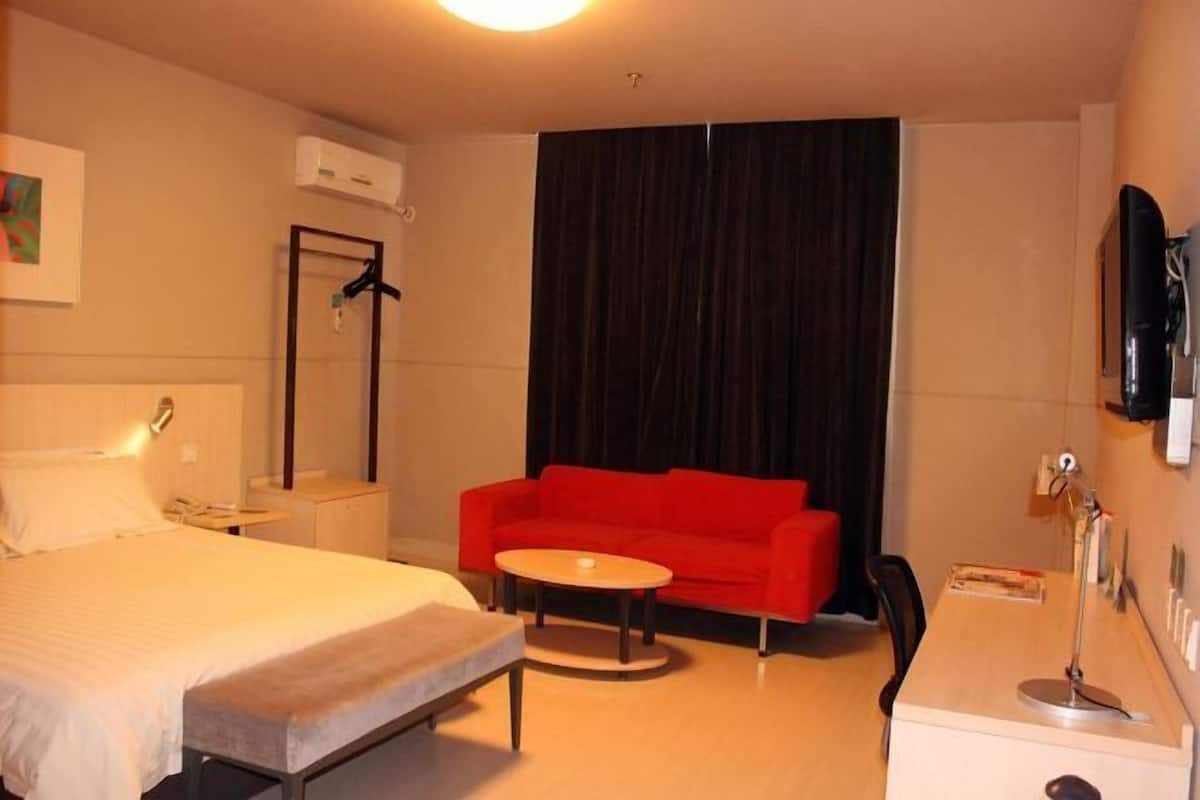 Ett hotellrum med en säng, en röd soffa, ett litet runt bord, ett skrivbord med en lampa och en svart gardin.