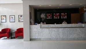 A hotel reception area with a marble counter, red chairs, framed artwork on the wall, and a receptionist.