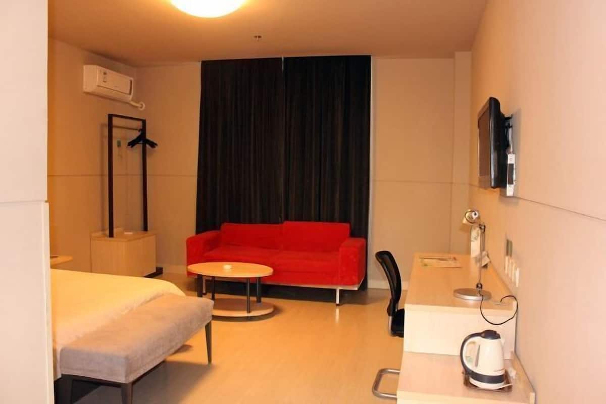 Ett hotellrum med en säng, en röd soffa, ett litet runt bord, ett skrivbord med en vattenkokare, en spegel och en luftkonditioneringsanläggning.
