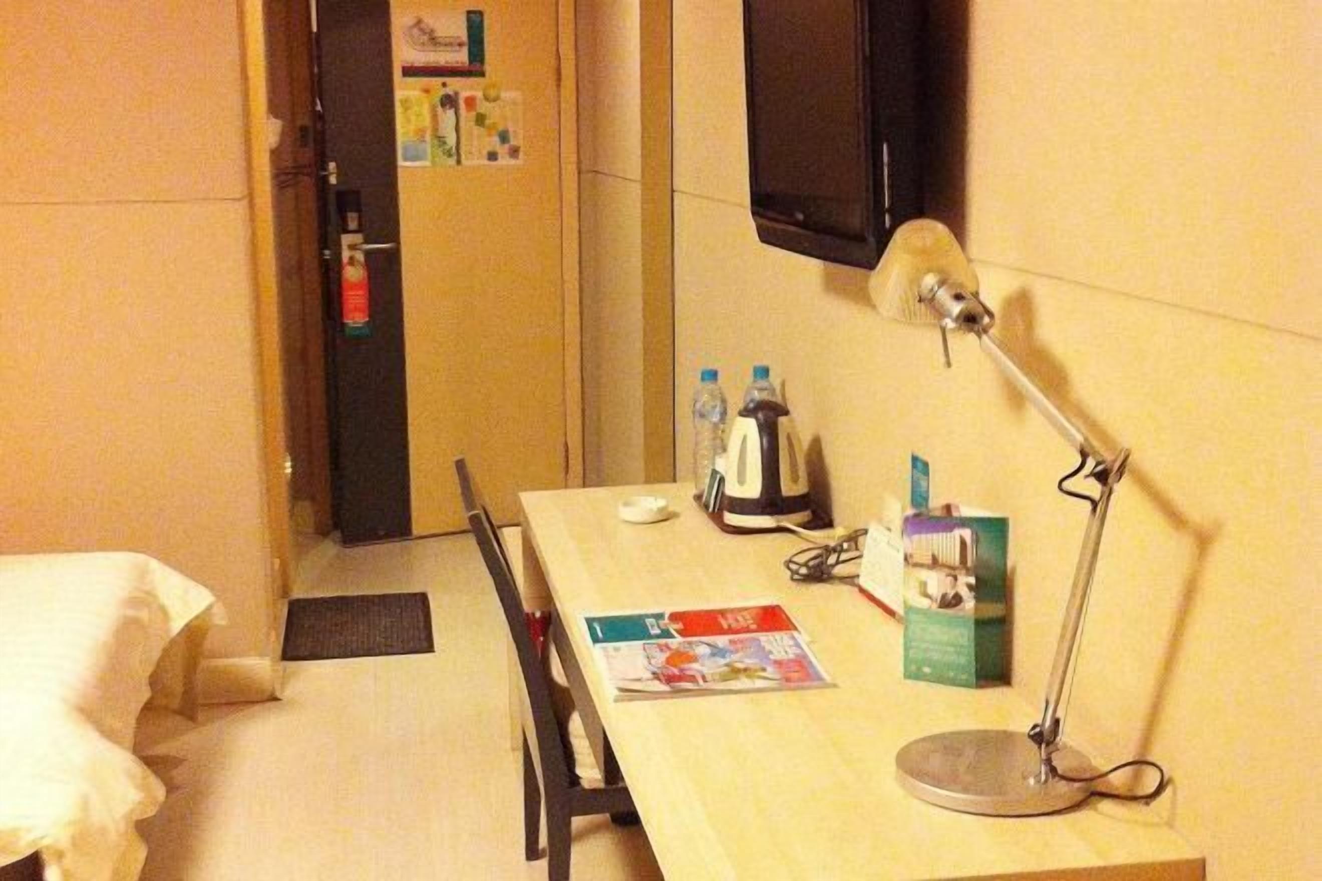 Quarto de hotel com escrivaninha de madeira, luminária, chaleira, garrafa de água, revista e porta com extintor de incêndio.