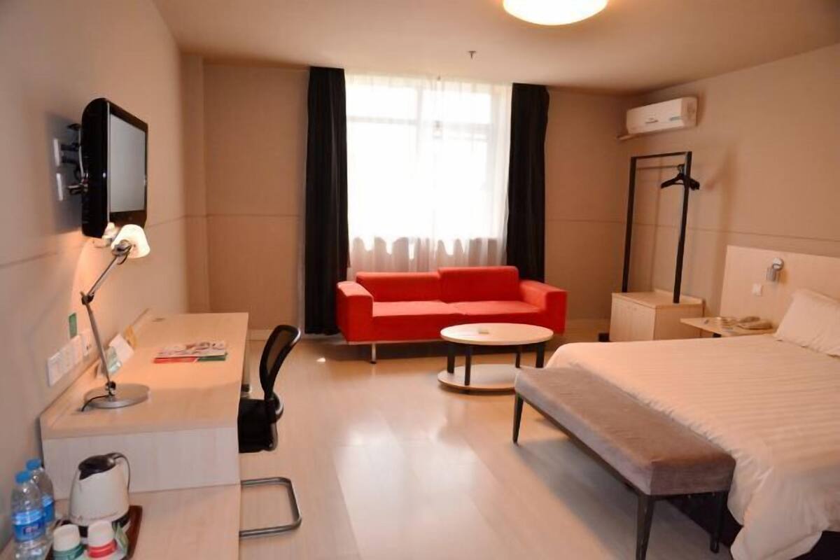 Ett hotellrum med en säng, ett skrivbord med en lampa, en röd soffa, ett litet runt bord och en tv som är monterad på väggen.