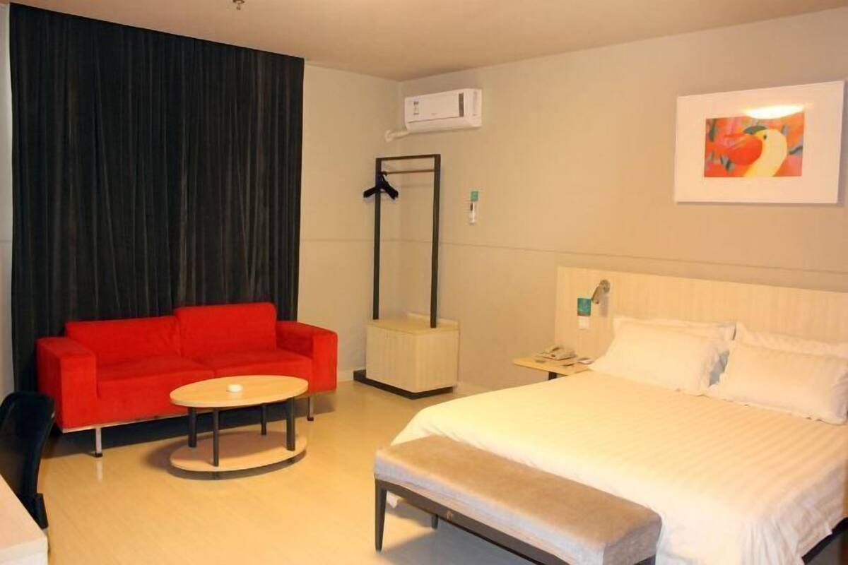 Ett hotellrum med en säng, en röd soffa, ett litet runt bord, en spegel och en väggmonterad luftkonditioneringsanläggning.