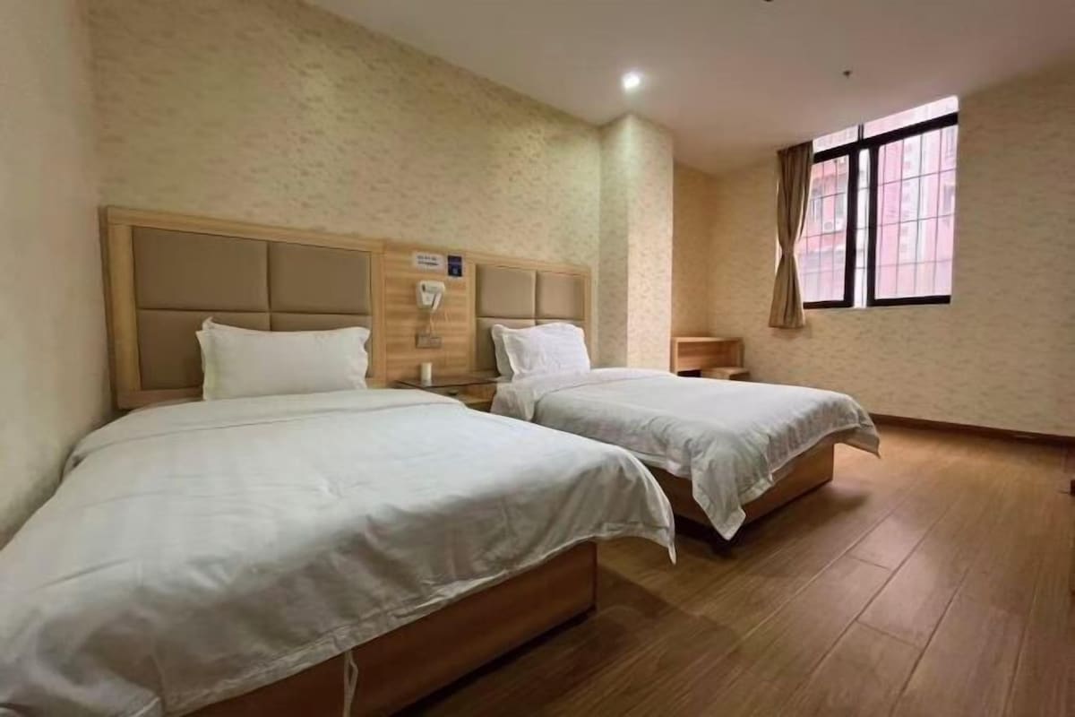 Habitación de hotel con dos camas, cabecero de madera, mesita de noche con lámpara y ventana con cortinas.