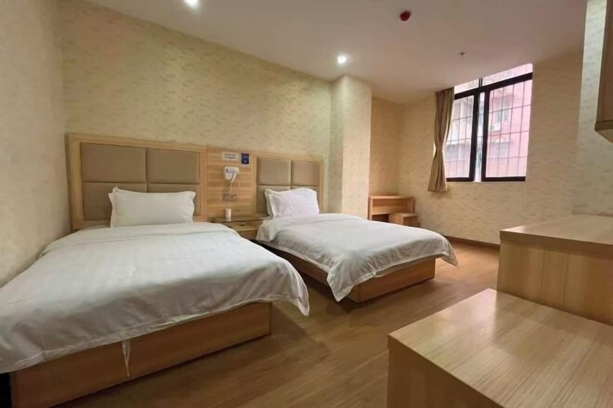 Une chambre d’hôtel avec deux lits, une tête de lit en bois, une table de chevet, une fenêtre avec des rideaux et un petit bureau en bois.