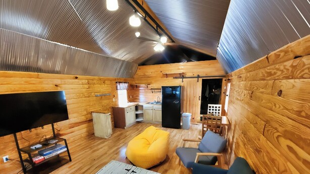 A cozy cabin interior with a wooden floor, a sofa, a yellow bean bag, a TV on a stand, and a kitchen area with a refrigerator.