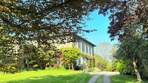 Ein Weg führt zu einem großzügigen Gebäude mit einer symmetrischen Fassade, umgeben von gepflegter Bepflanzung und Bäumen.