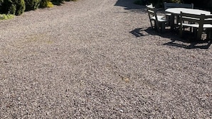 Ein Kiesweg führt zu einem Holztisch und -stühlen, umgeben von dichtem Grün und hohen Bäumen.