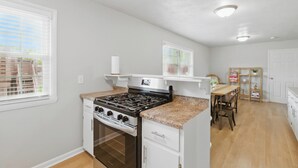 Una cocina compacta con estufa a gas, horno y muebles blancos. Hay un comedor con mesa y sillas de madera.