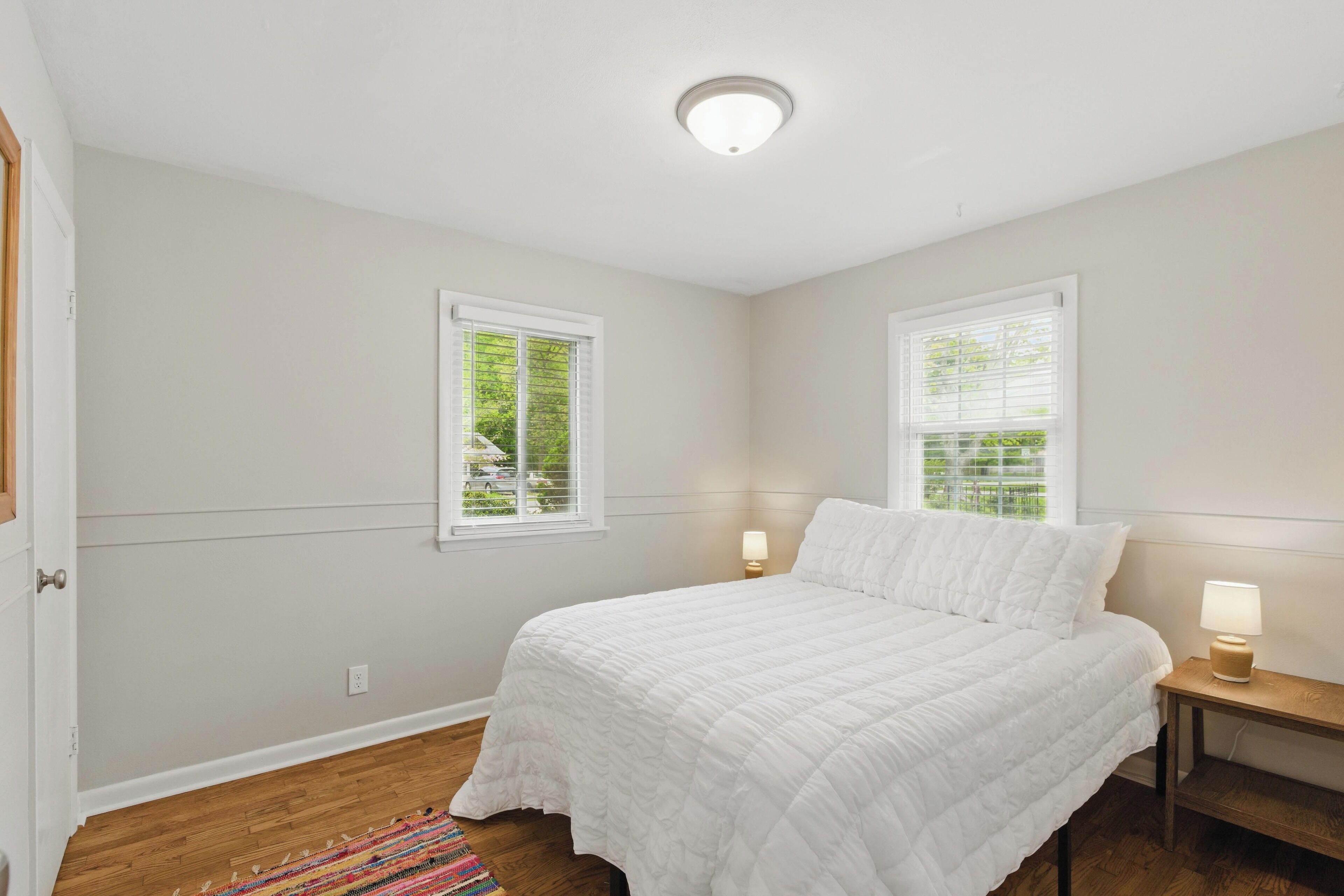 Un dormitorio con una cama, dos ventanas, una mesita de noche con lámpara y suelo de madera.