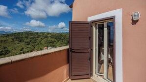 A balcony with a view of green hills and mountains, a brown door, and a window with a light fixture.