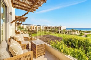 A balcony with wicker furniture, a glass balustrade, and a view of a resort complex and the sea.