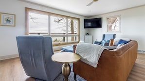 A living room with a brown leather sofa, a blue armchair, a small round table, and a ceiling fan. - Lakefront Cottage with Hot Tub & Beach Access (Fennville)