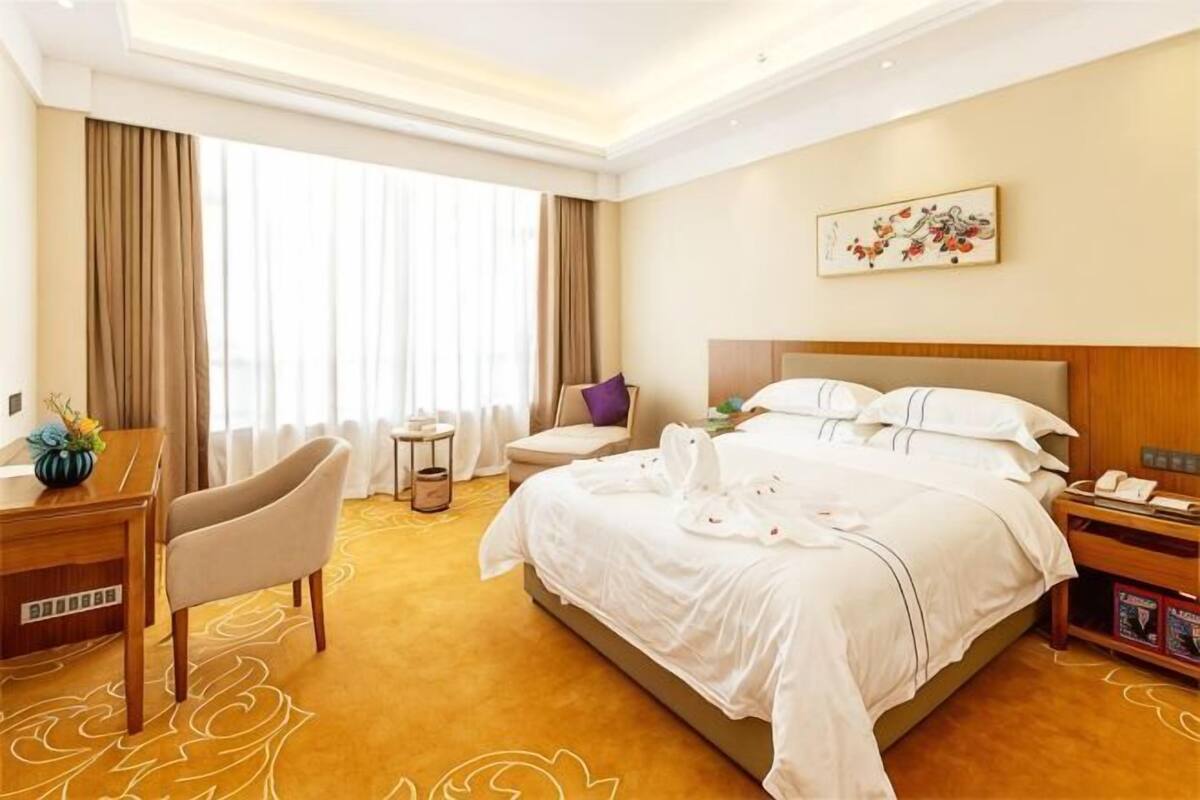 Quarto de hotel com uma cama grande, uma escrivaninha, uma cadeira e uma janela com cortinas.