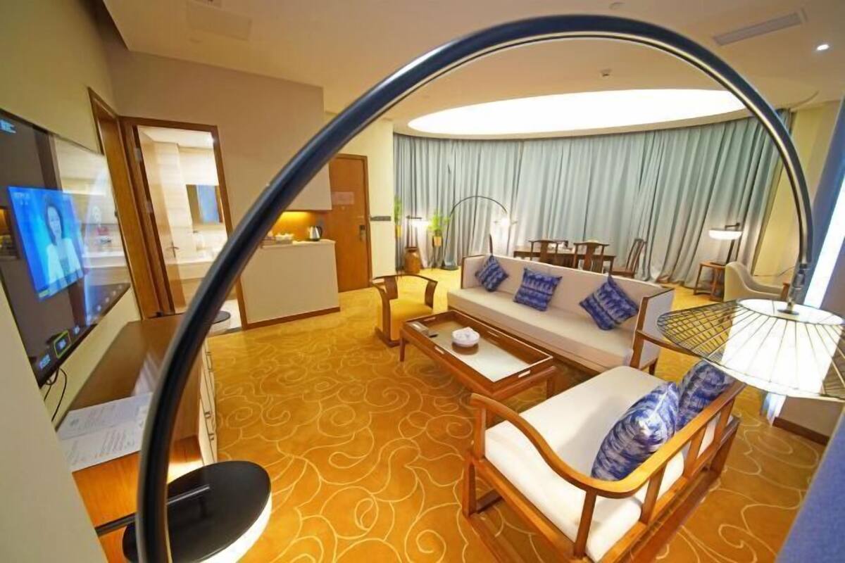 Quarto de hotel com sofá, poltronas, mesa de centro, televisão e área de jantar com mesa e cadeiras.