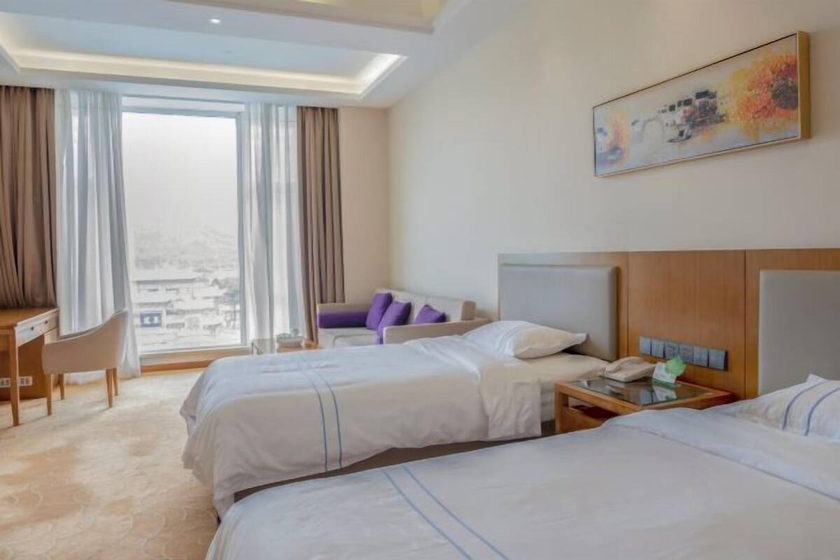 Quarto de hotel com duas camas, uma escrivaninha, uma cadeira e uma janela grande com cortinas.